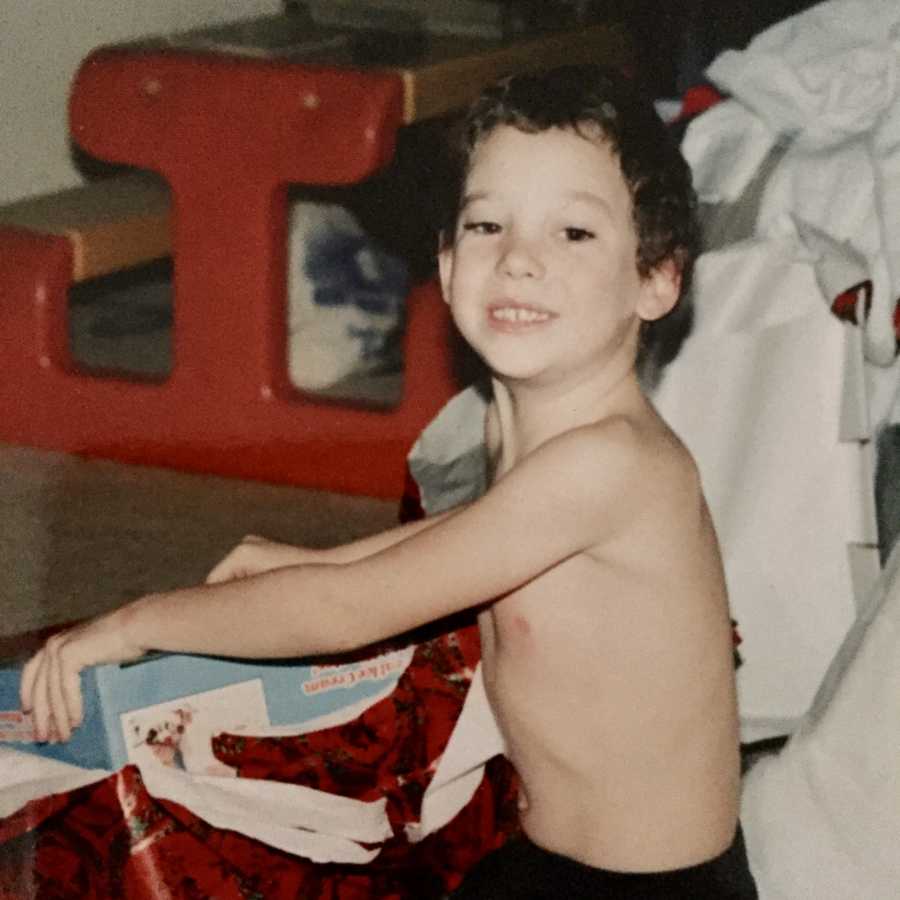 Young boy without a shirt on smiles as he opens present 
