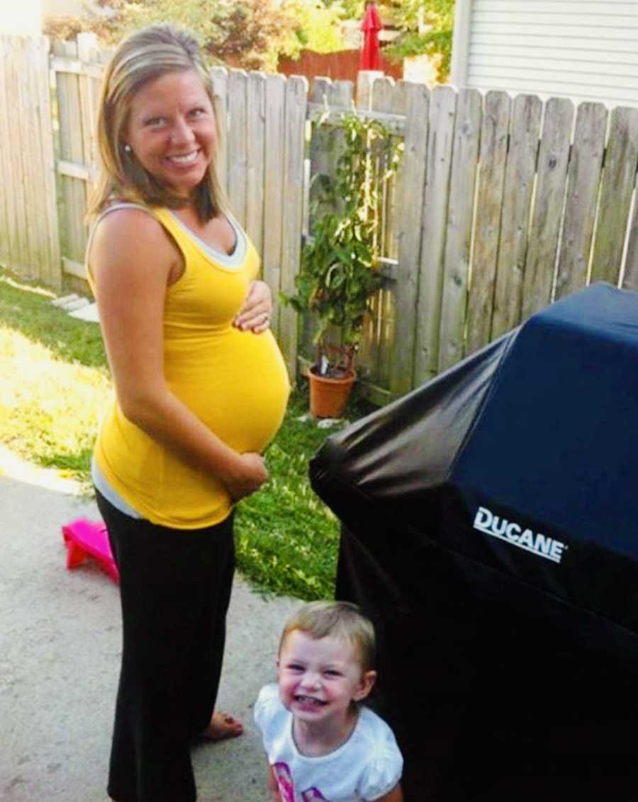 Pregnant woman smiles outside beside her daughter and covered grill
