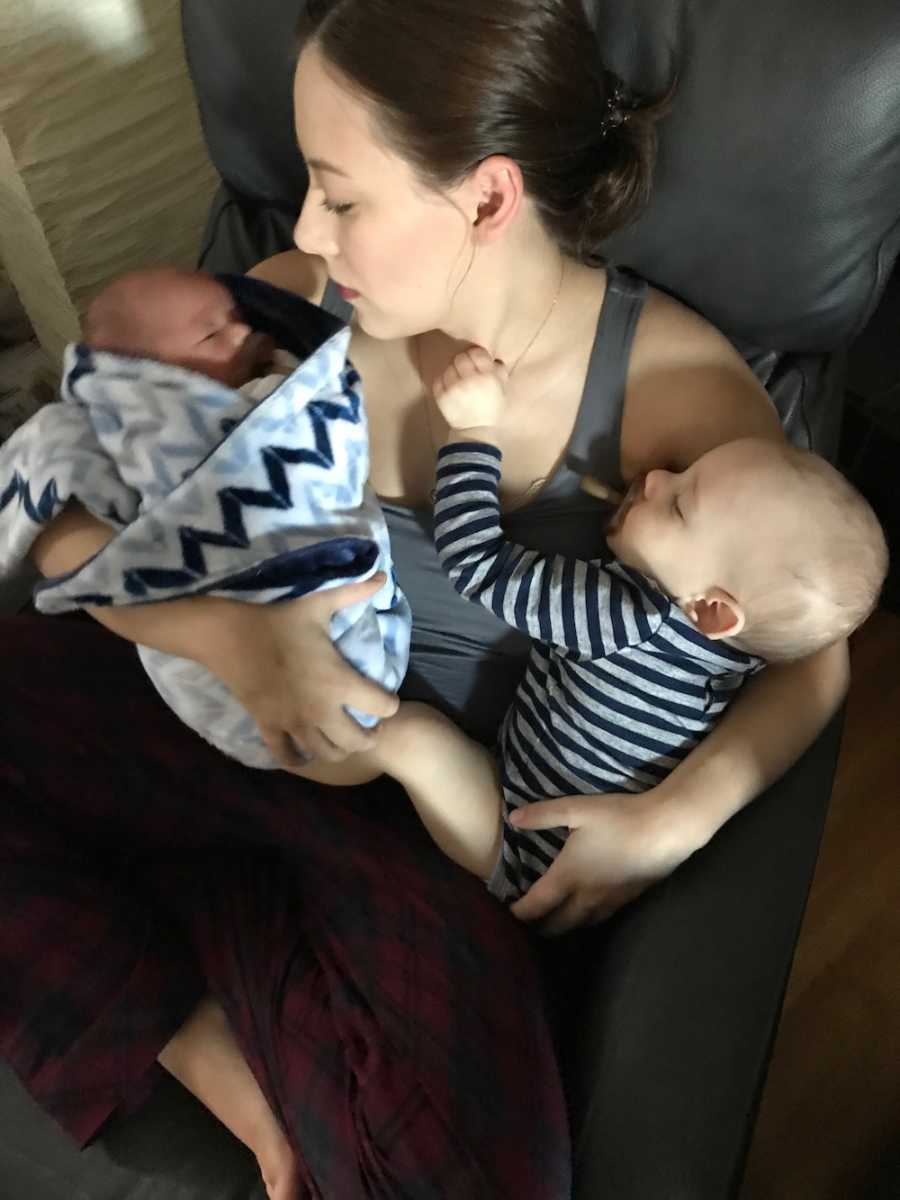 Woman sits in chair with first born asleep in one arm and newborn asleep. in other