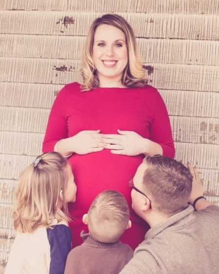 Pregnant woman smiles holding her stomach while her husband and his two kids kiss her stomach