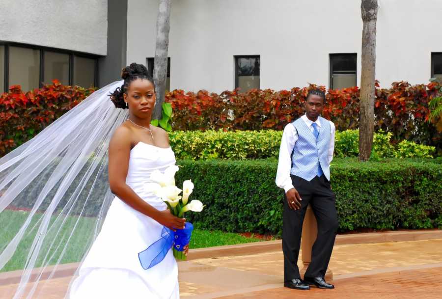 Bride stands holding flowers and veil flowing in wind while husband stands in background watching