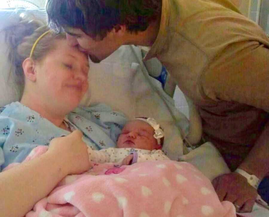 Woman lies in hospital bed with newborn in her arms while husband leans over to kiss her on forehead