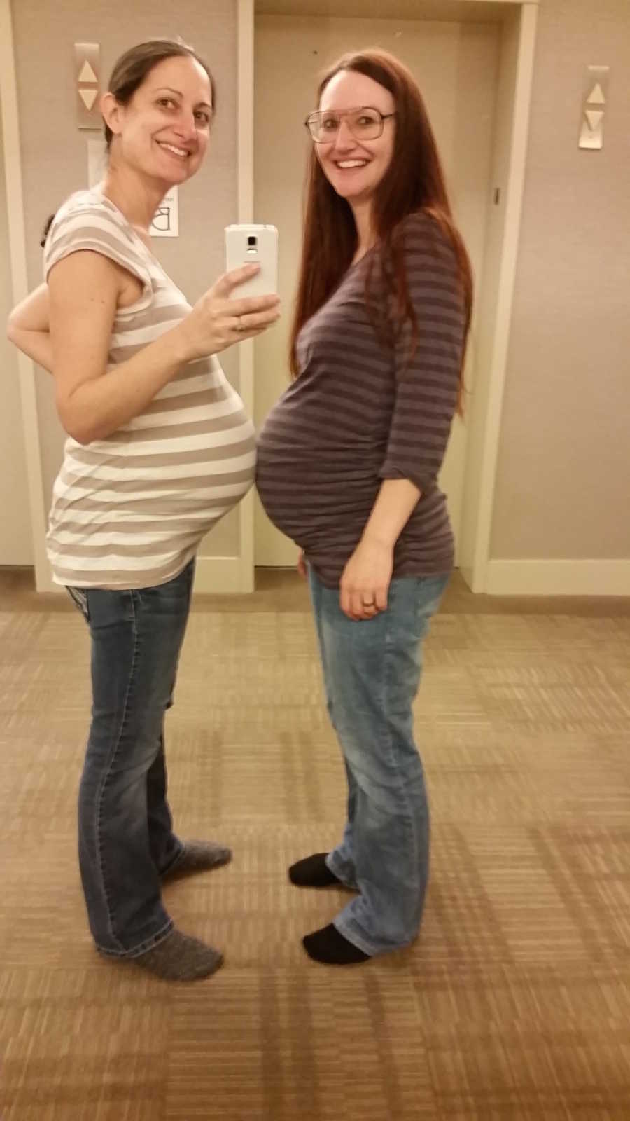 Pregnant woman smiles with stomach touching pregnant friends stomach in mirror selfie