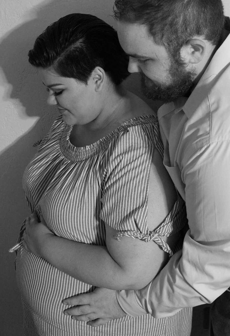 Pregnant woman stands holding her stomach while her husband stands behind her holding her side