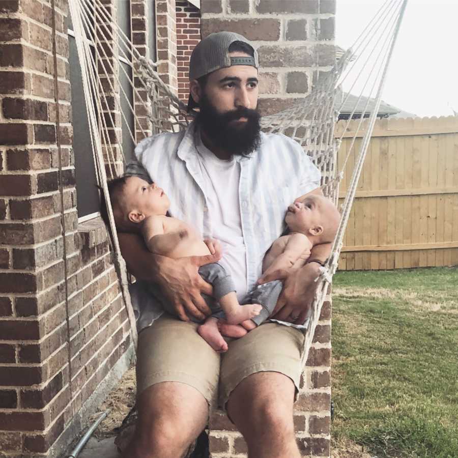 Stay at home father sits on outdoor swing holding twin boys