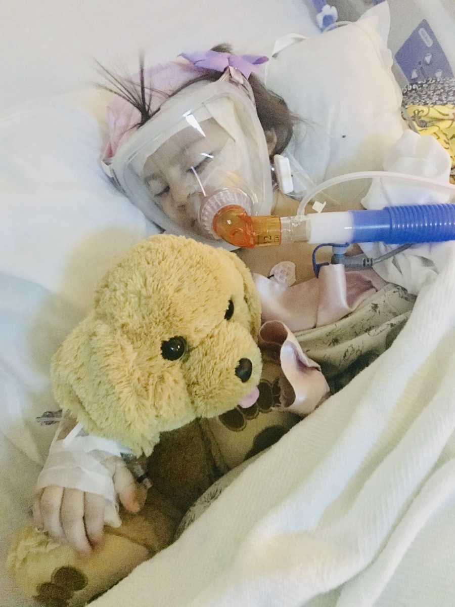 Little girl with acute flaccid myelitis lays asleep in hospital with oxygen mask and her arm around stuffed animal