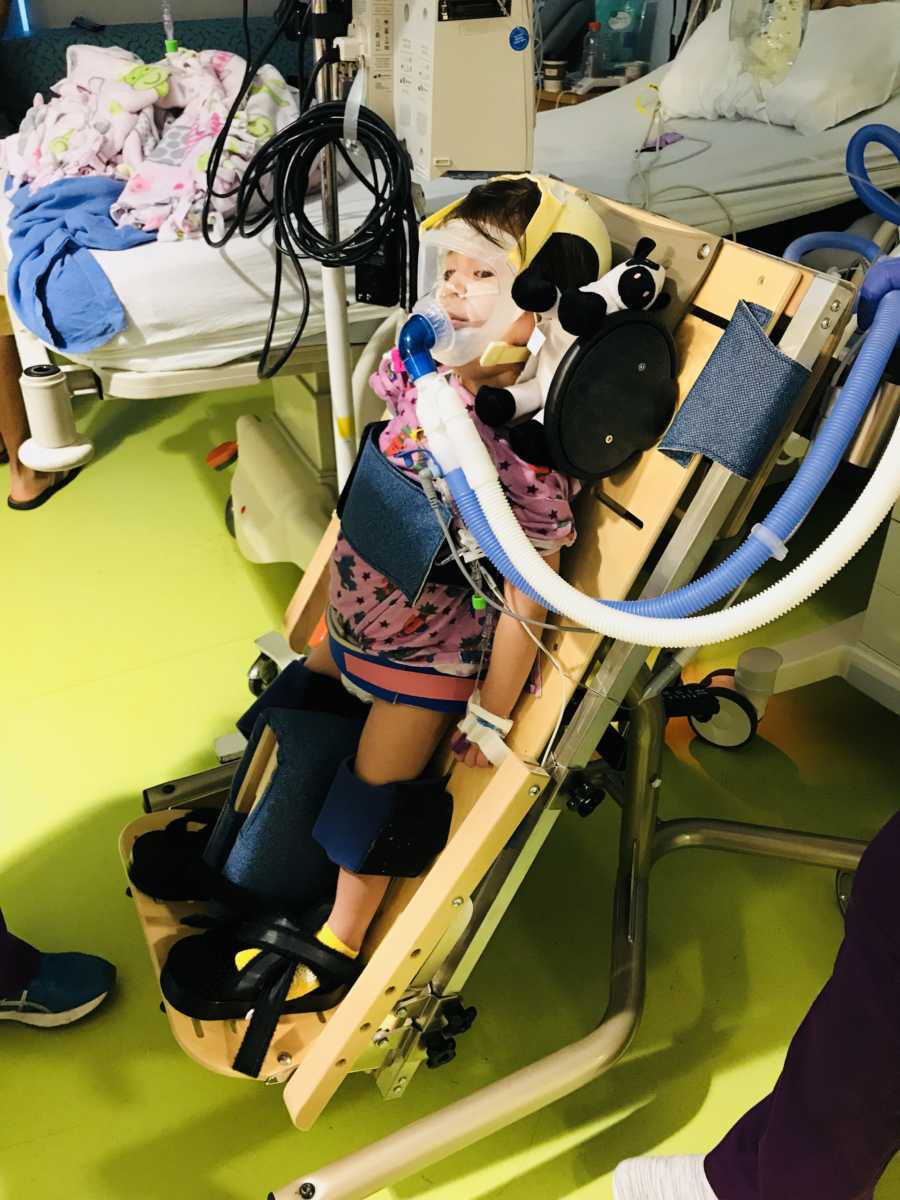Little girl with acute flaccid myelitis stands strapped to wooden board in hospital room with oxygen mask