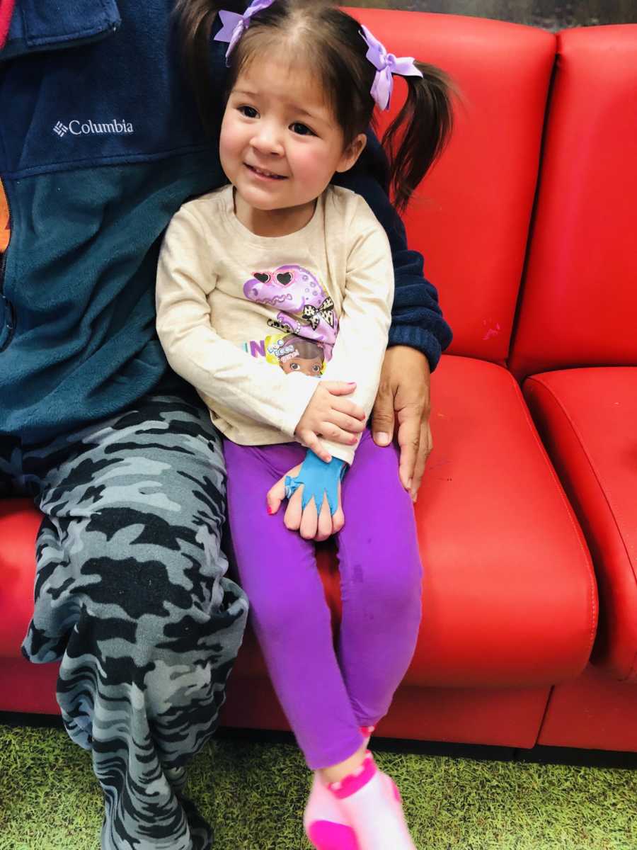 Little girl with acute flaccid myelitis sitting on red couch with father's arm wrapped around her