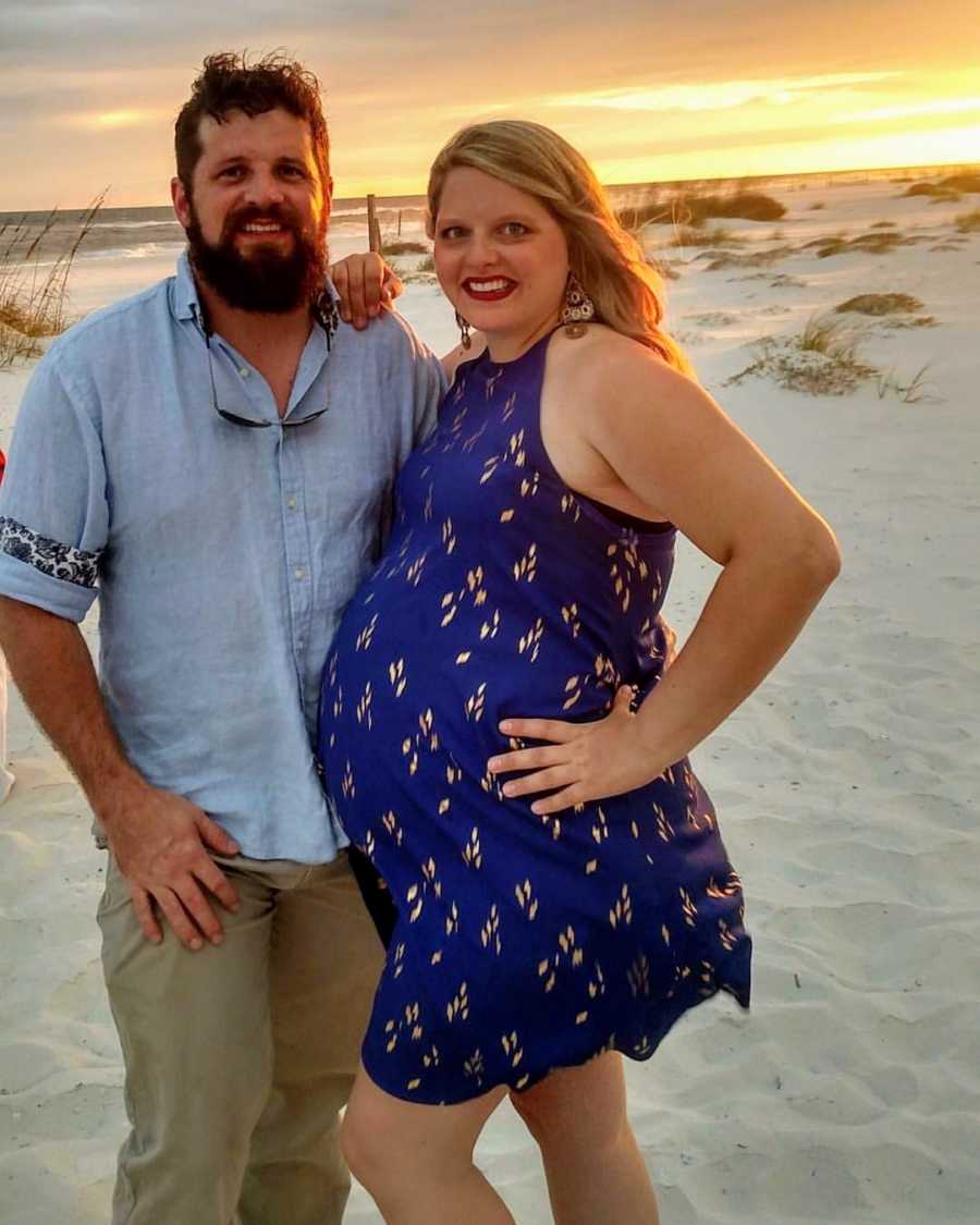 Pregnant woman stands arm in arm with husband on beach at sunset
