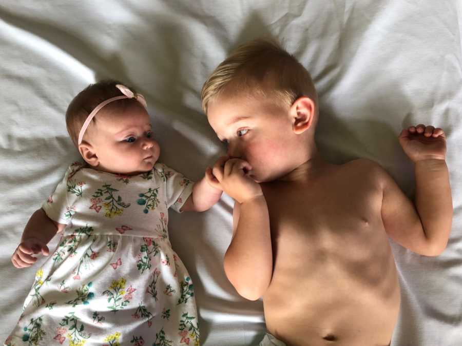 Infant laying shirtless in bed beside baby sister kissing her hand