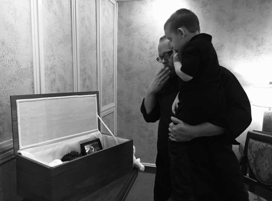 Father holds son as they blow a kiss to deceased baby in open casket 
