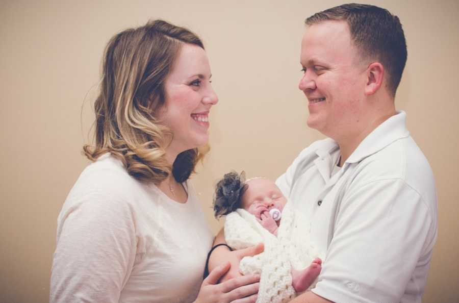 Husband and wife smile looking into each others eye while husband holds newborn baby girl