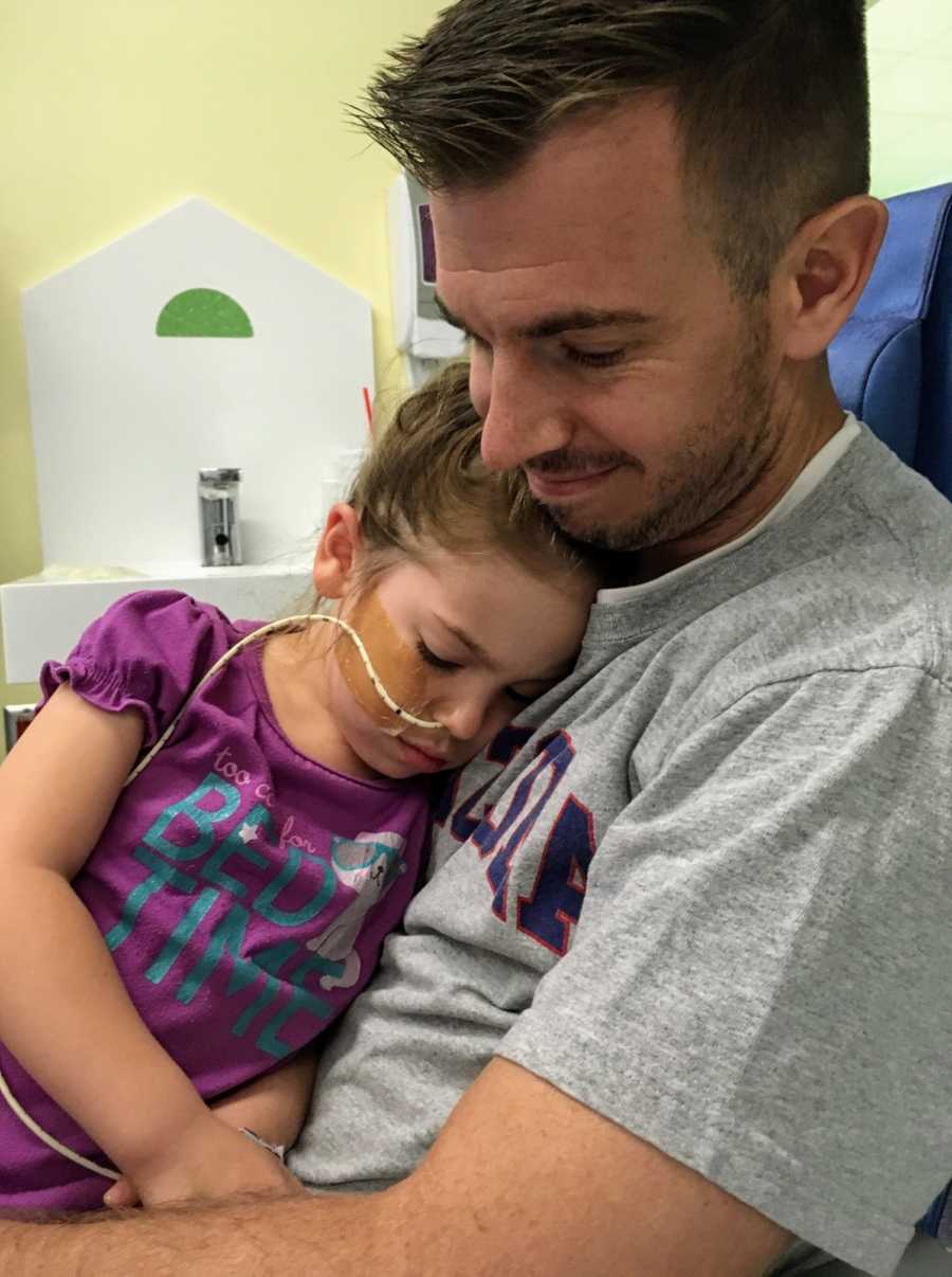Father sits in chair in doctor's office with sick daughter in his lap