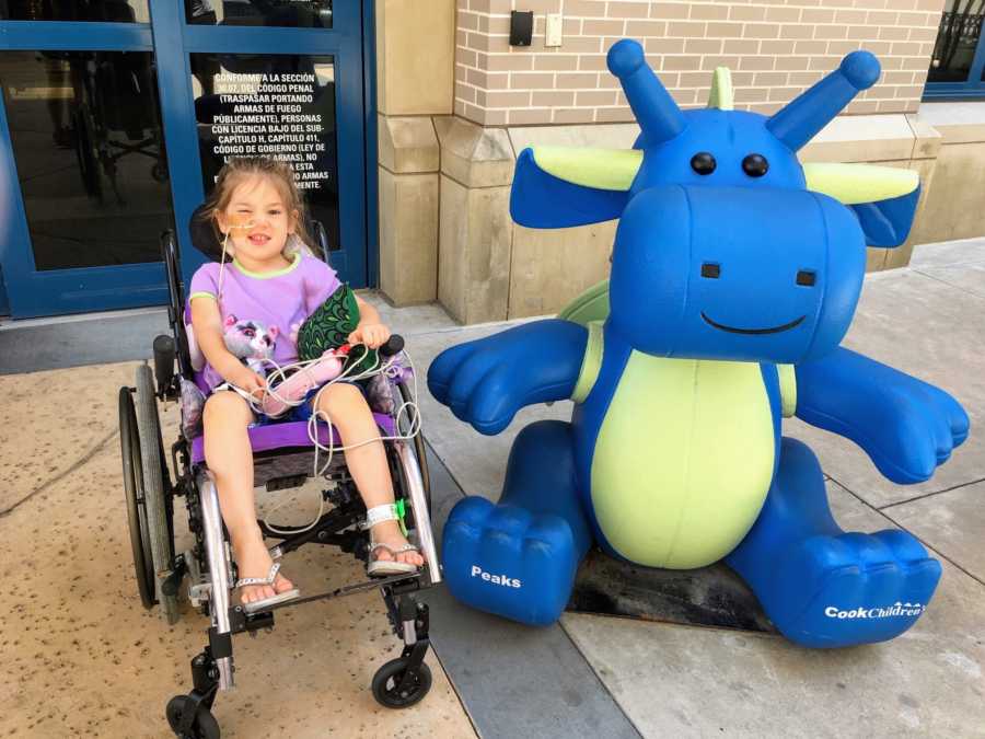 Young girl with polio-like symptoms sits in wheelchair outside hospital beside dinosaur statue