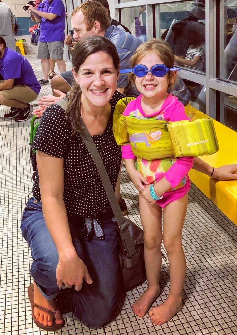 Mother crouches down next to daughter on pool deck wearing floaties and goggles