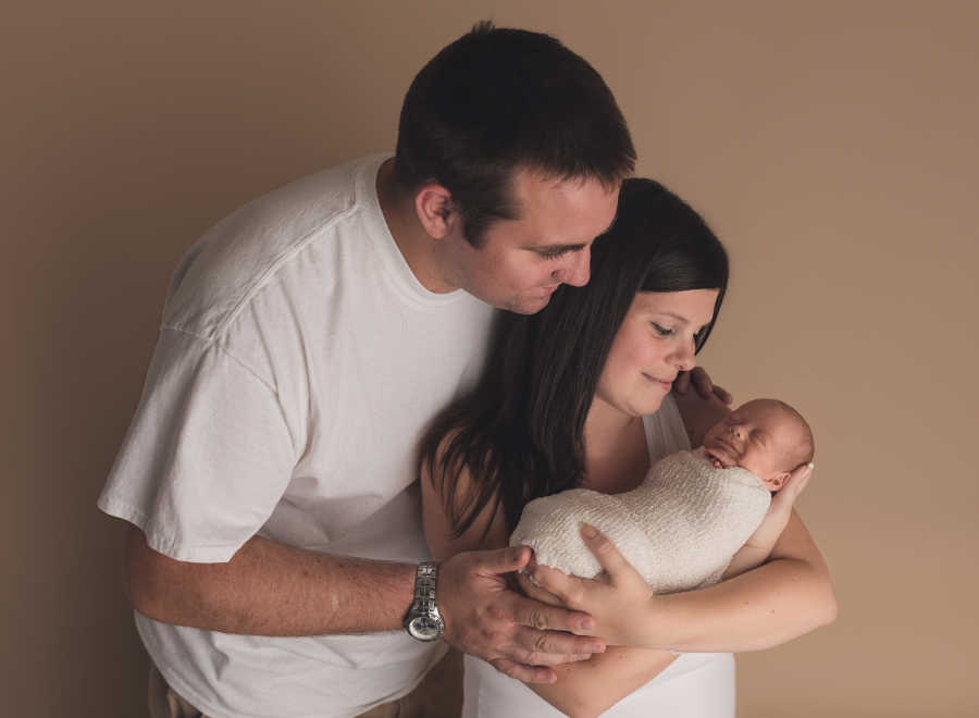 Mother holds her daughter swaddled in her arms while husband stands beside her