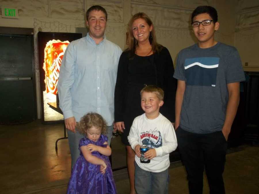 Husband and wife stand beside their teen foster child with their biological kids in front of them