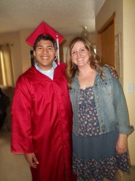 Woman stands smiling beside foster son who stands in red cap and gown