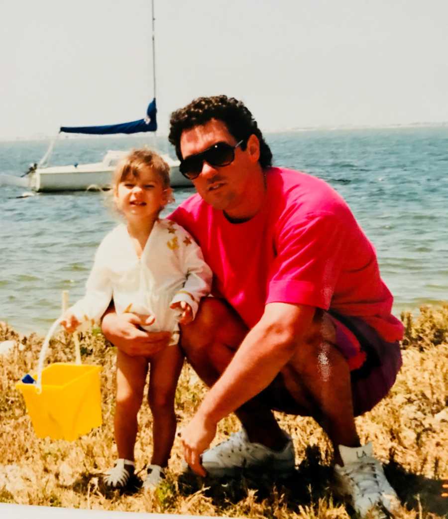 Man squatting beside young daughter who holds bucket beside body of water with sailboat