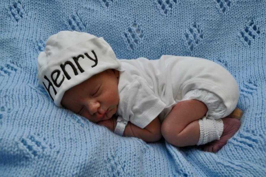 Newborn who almost died during birth lays on blue blanket in white onesie and hat that says, "Henry"