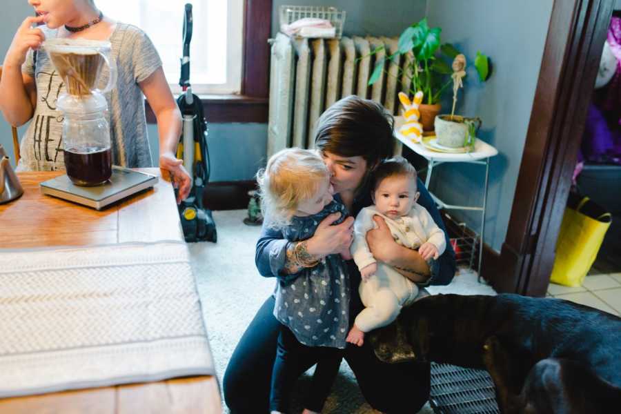 Woman squats to ground kissing foster child while holding foster babu while older foster child stands making coffee