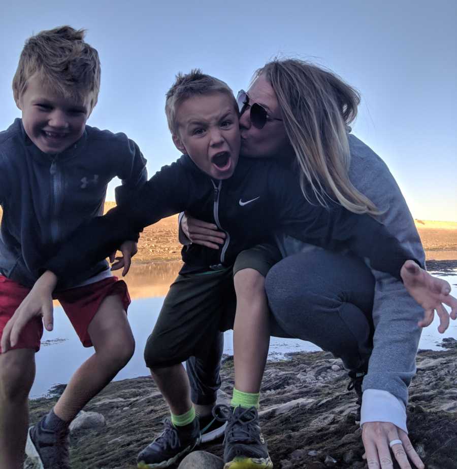 Woman with Sarcoma Cancer kneels on ground beside kissing cheek of one of her sons beside other son