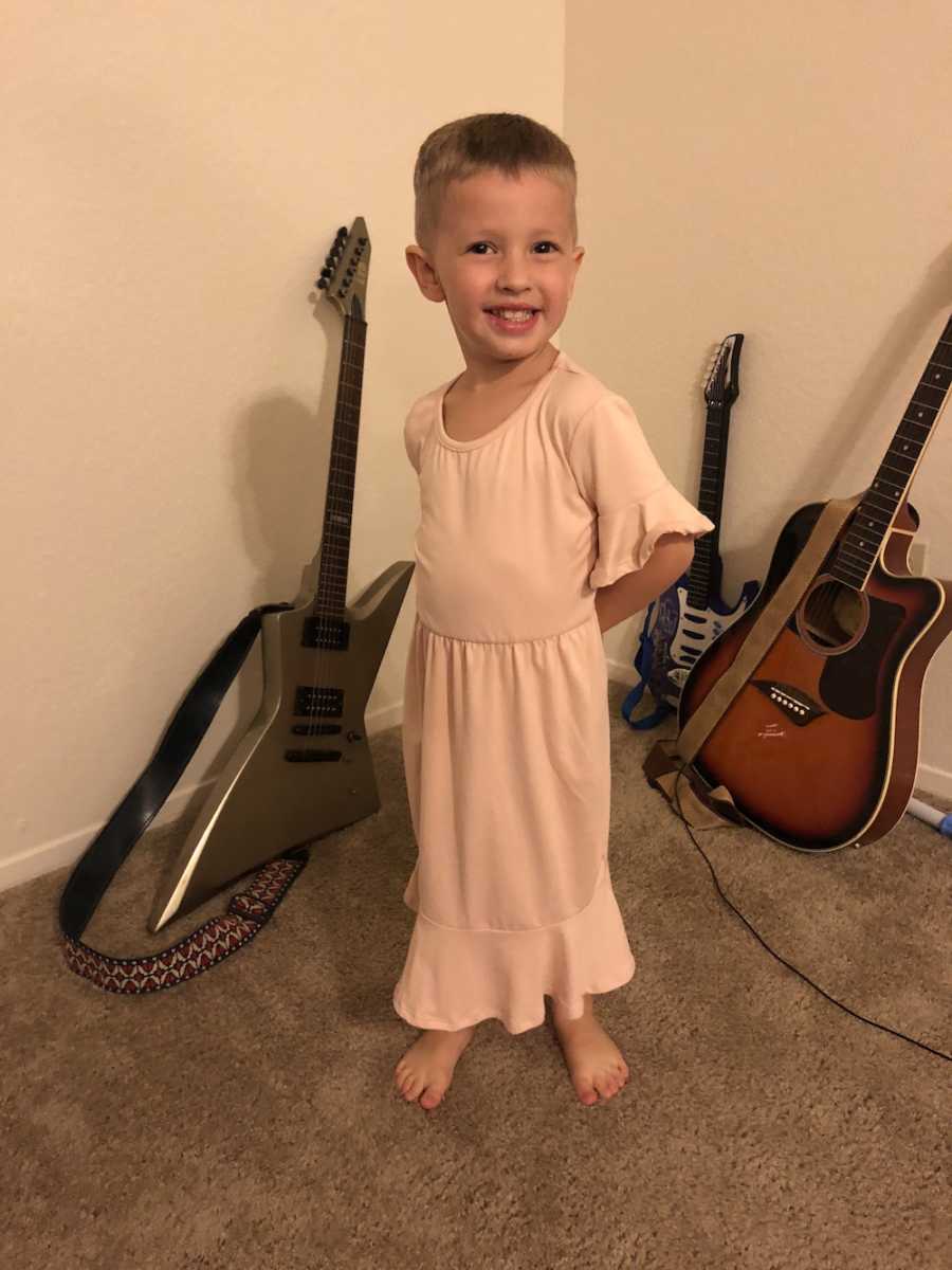 Little boy stands smiling wearing pink dress in front of guitars resting against the wall