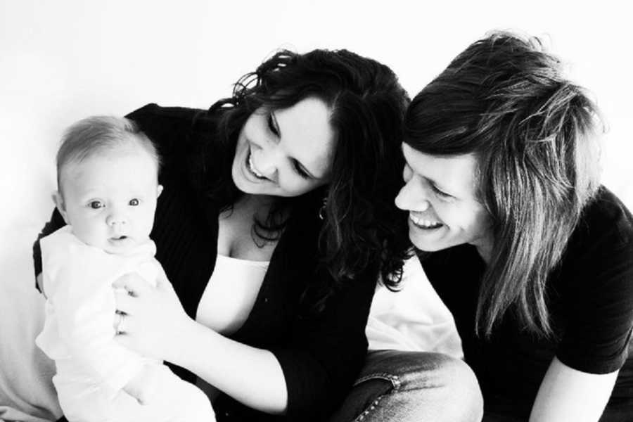 Mother and father sit on ground smiling at baby that is sitting on wife's lap in photoshoot