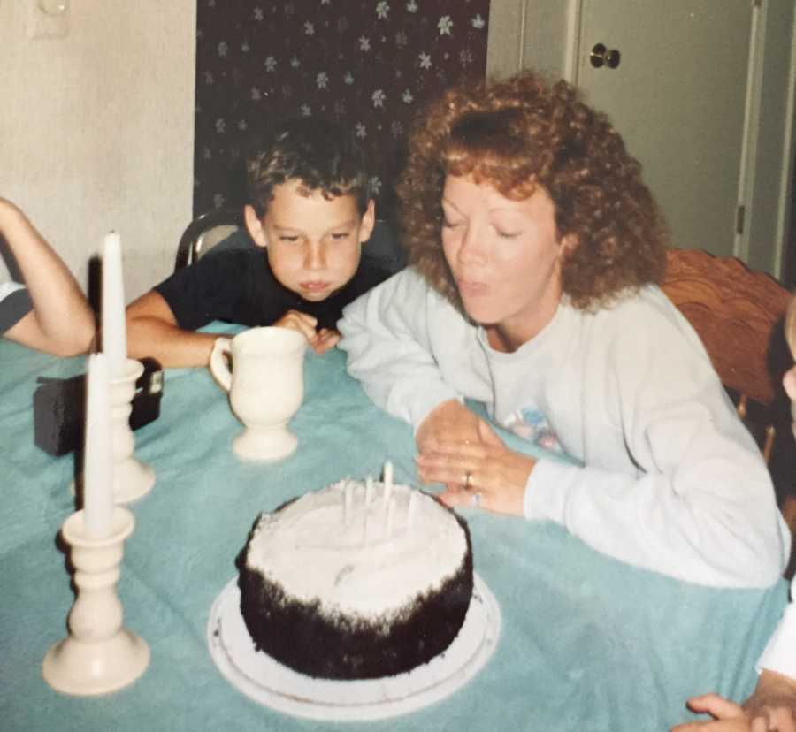 Mother sits at kitchen table blowing out candles in her cake with son by her side