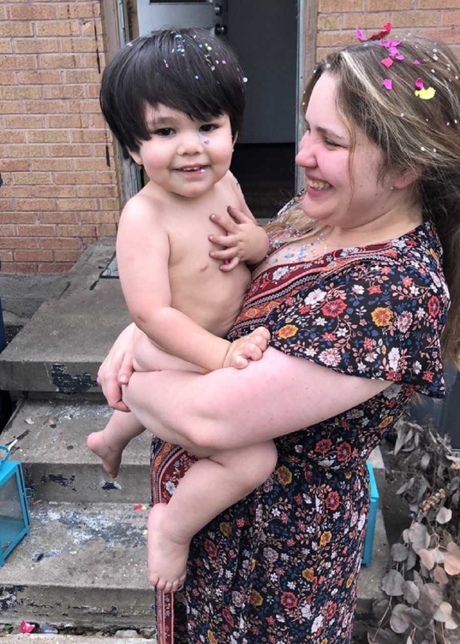 Mother with confetti in her hair stands holding toddler son who is also covered in confetti