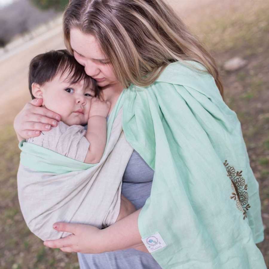 Mother stands outside holding son swaddled to her chest