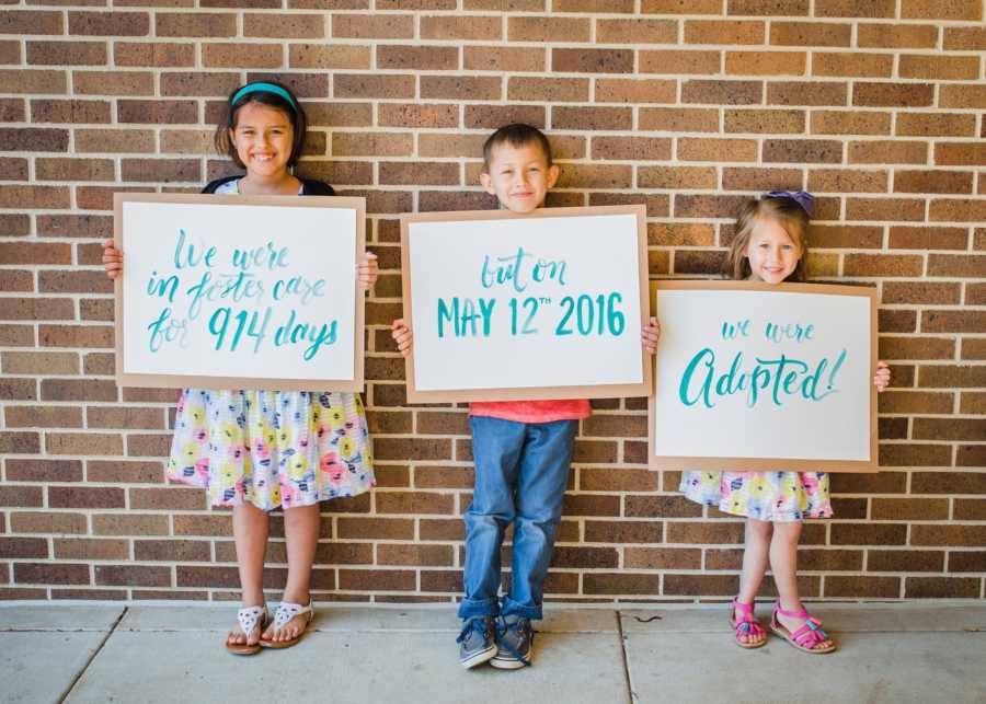 Three foster children stand against brick wall holding signs announcing their adoption