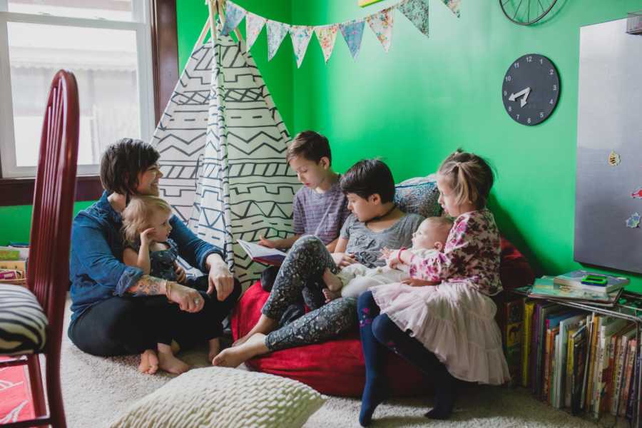 Woman sits on ground with foster baby in lap beside four other foster kids reading a book