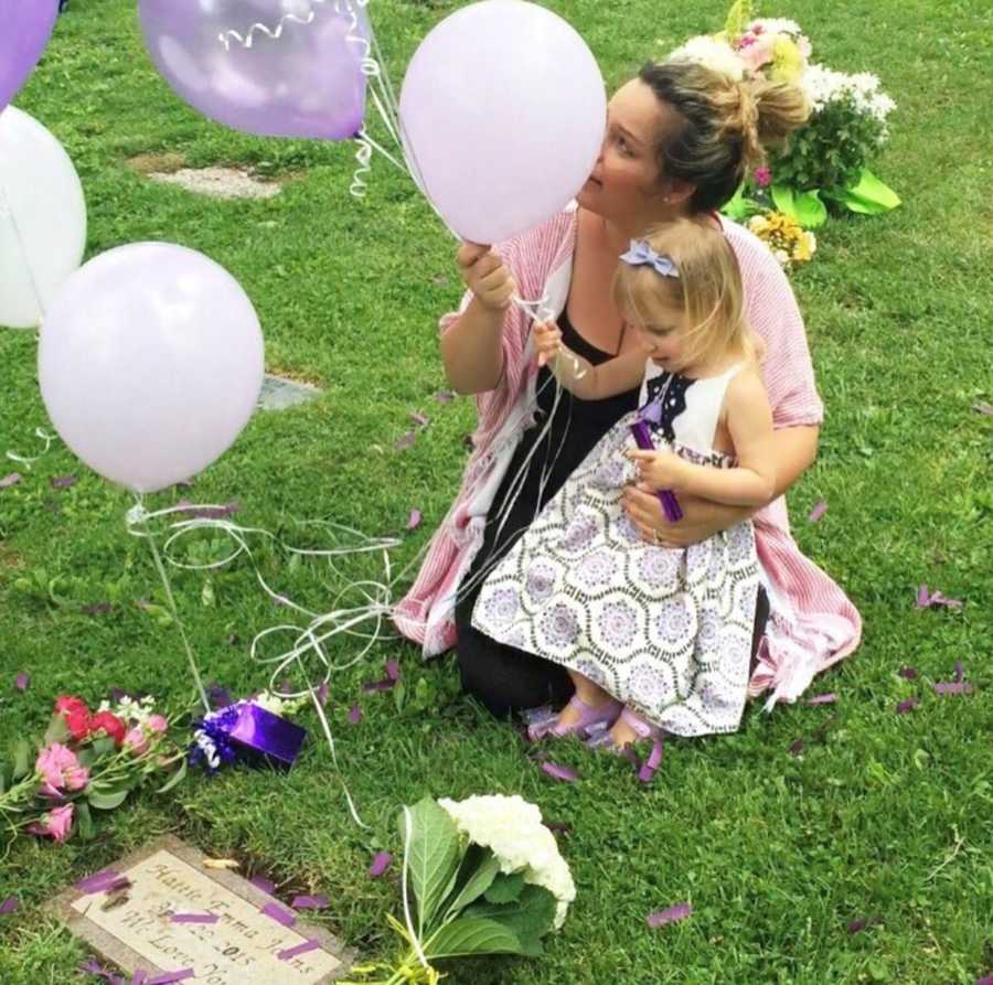 Mother sits on grass with daughter in her lap as they hold balloons in front of deceased daughter's grave