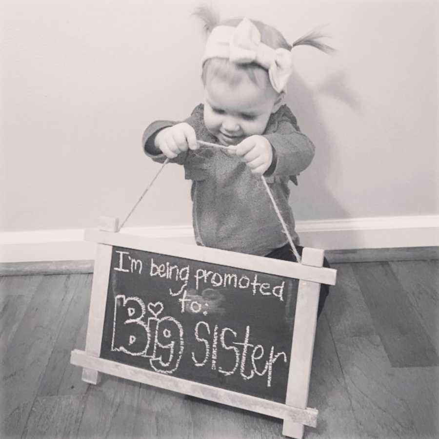 Little girl stands holding sign that says, I'm being promoted to big sister