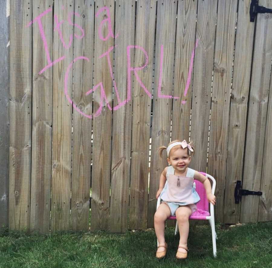 Little girl sitting in small pink chair in front of wooden fence that has "It's a girl" painted on it in pink