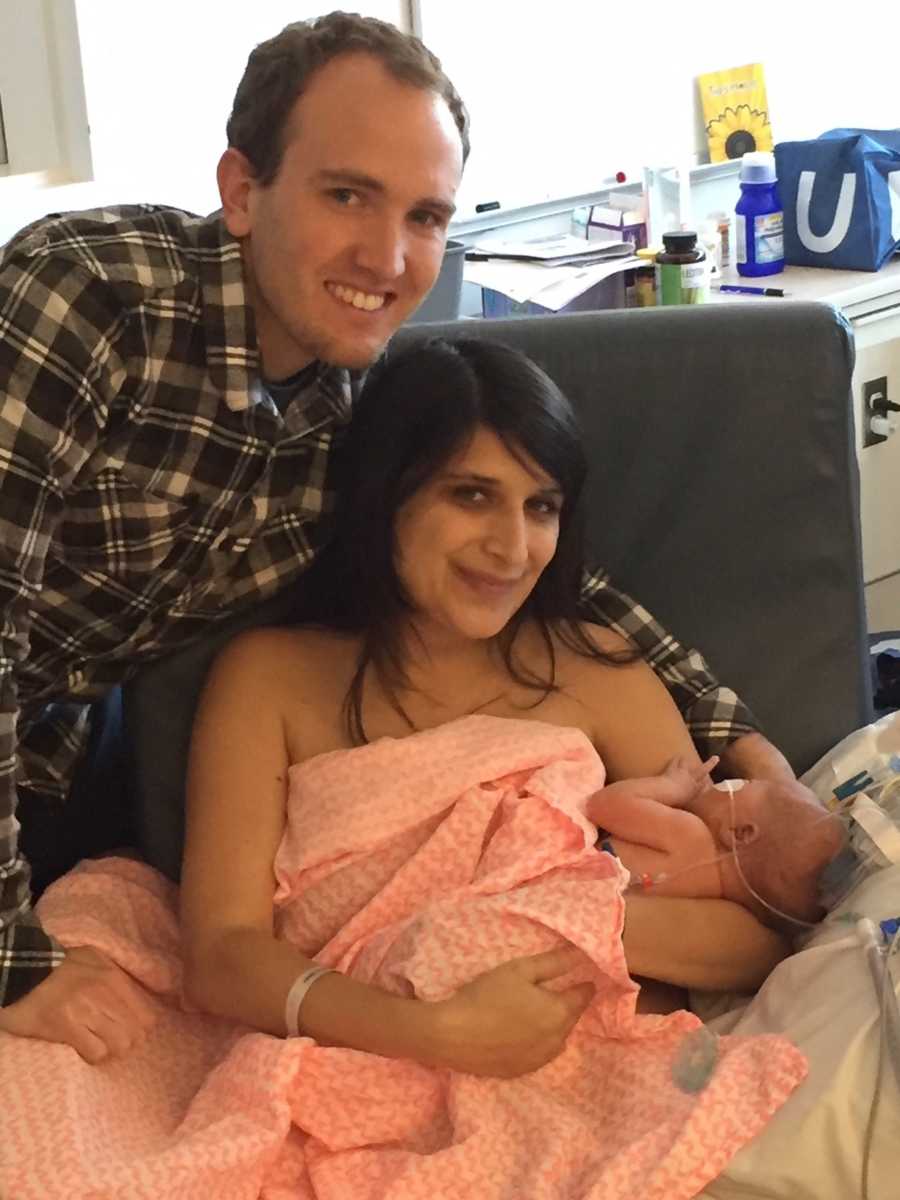 Mother sits in chair covered in blanket holding newborn in her arms with husband standing beside them