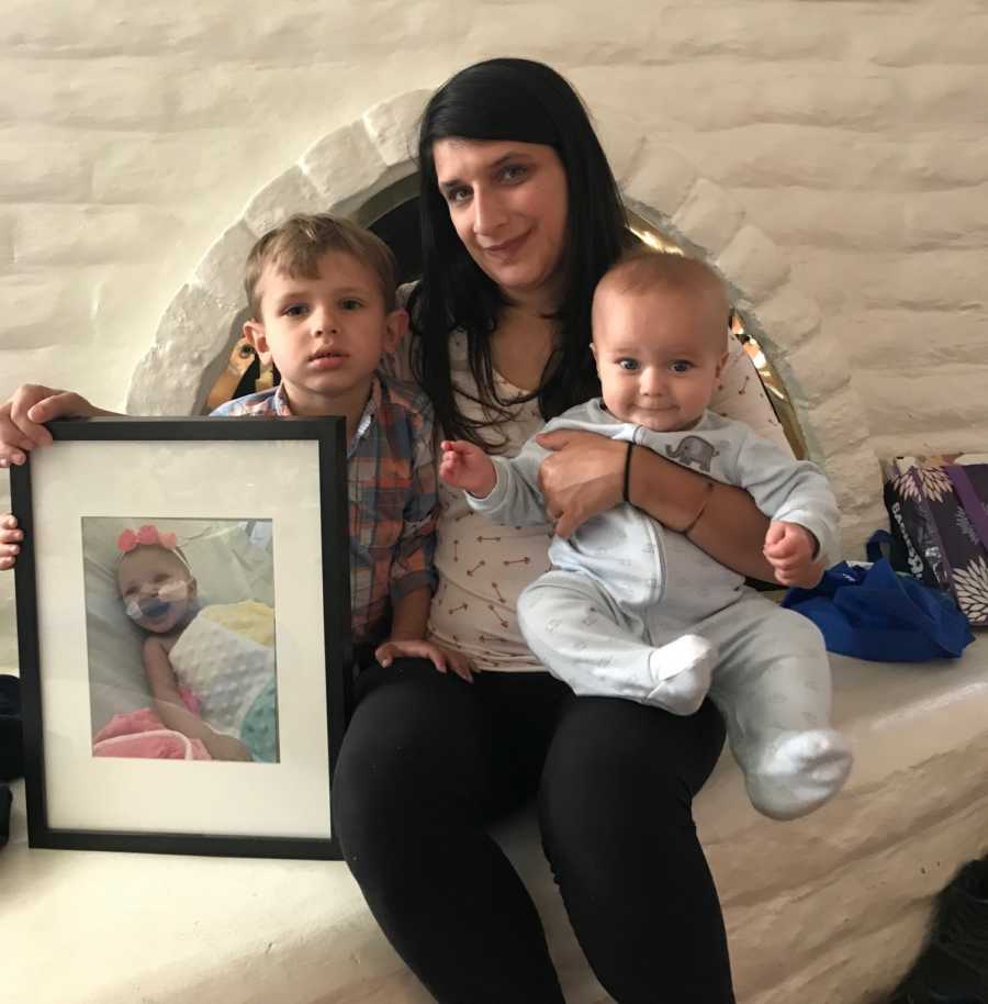 Mother sitting in front of fire place with baby on her lap, her son beside her, and picture frame of deceased daughter