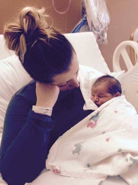 Woman sits in hospital bed with head resting on her first as she looks at newborn sleeping in her arm