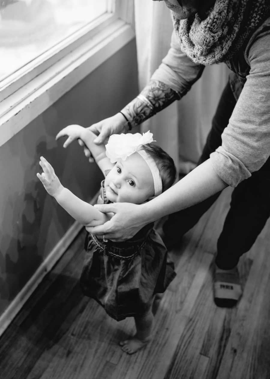 Baby in foster care standing beside window whose foster mother is holding her arms