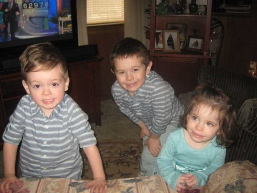 Young boy stands smiling behind his moms' two foster children who are leaning against couch