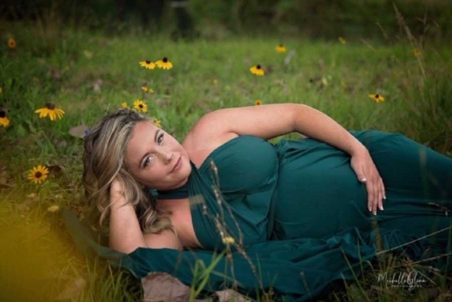 Pregnant woman lays on her side in teal gown beside sunflowers for photoshoot