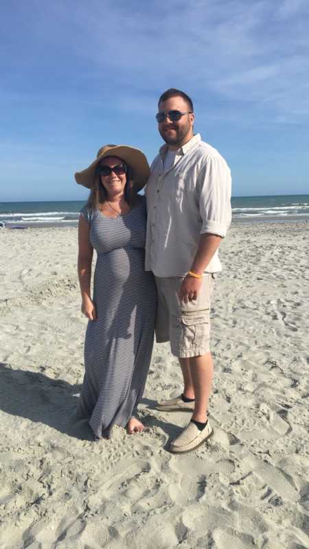 Pregnant woman stands smiling beside husband on beach