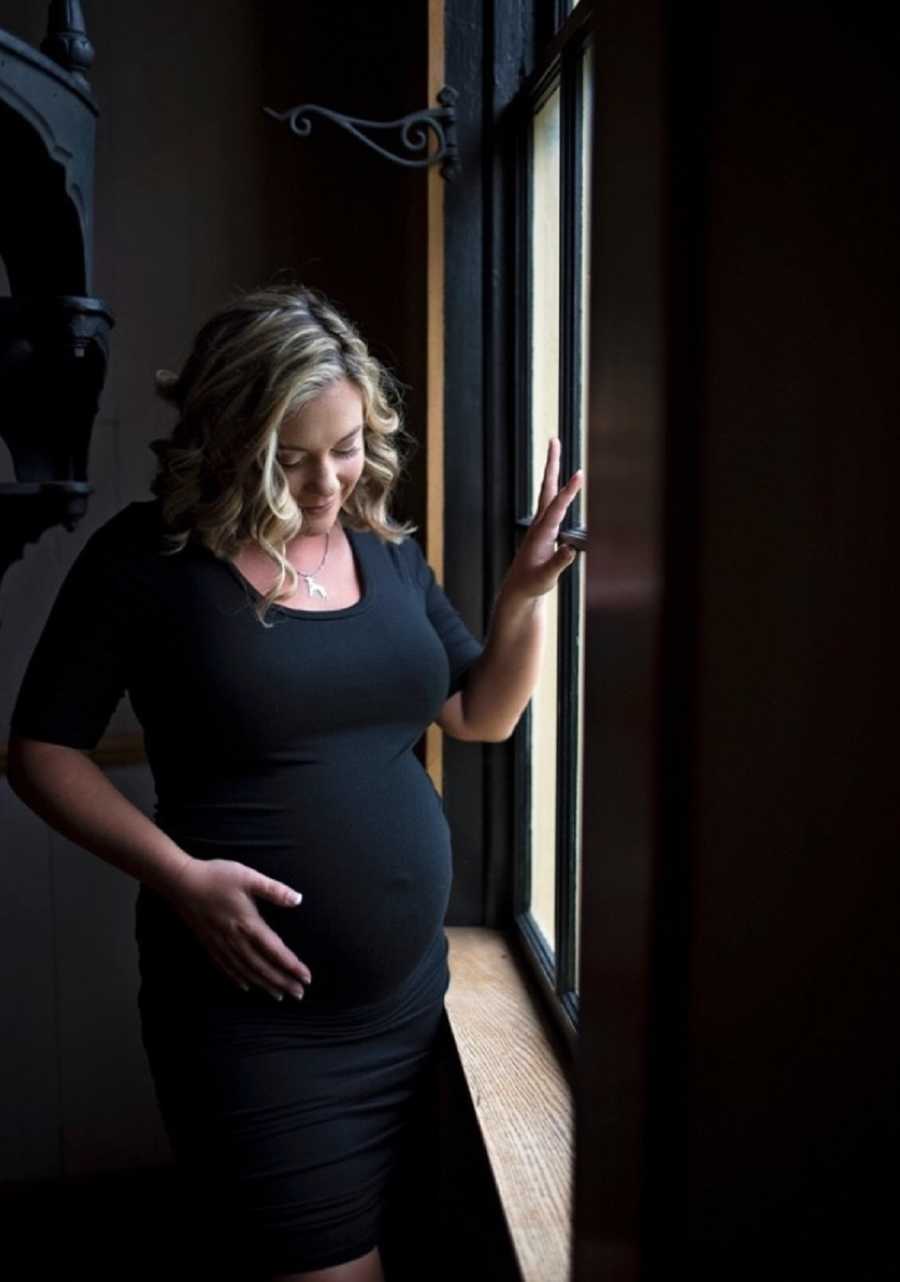 Pregnant woman stands holding one hand on stomach and other on window