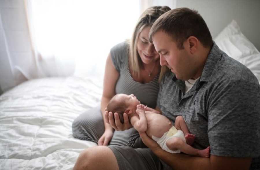 Husband and wife sit on bed while husband holds newborn that is finally home from NICU