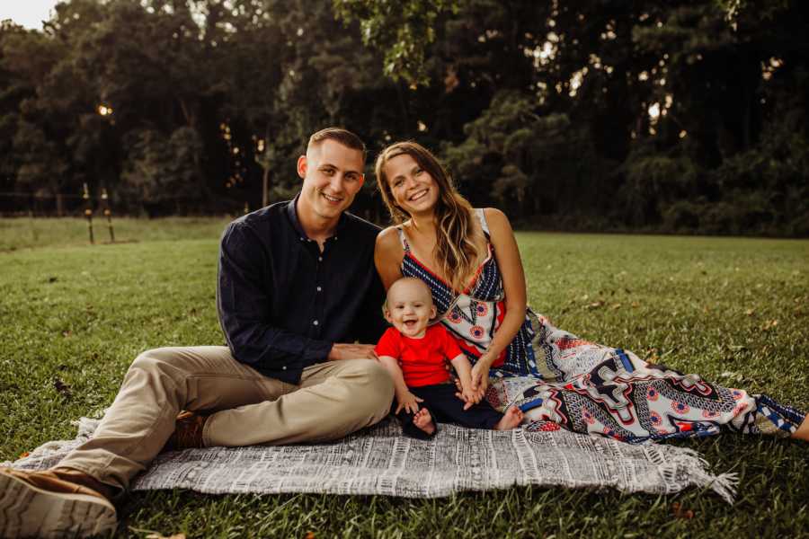 Husband and wife sit on blanket outside with infant son sitting between them