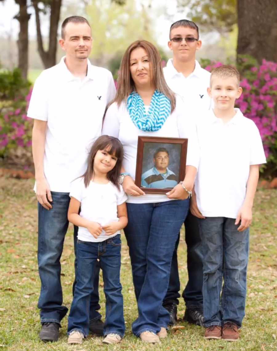 Woman stands holding picture of deceased brother beside three children in husband in family photo