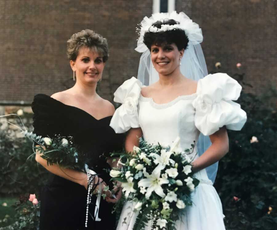 Bride stands holding bouquet of white flowers beside woman in black dress