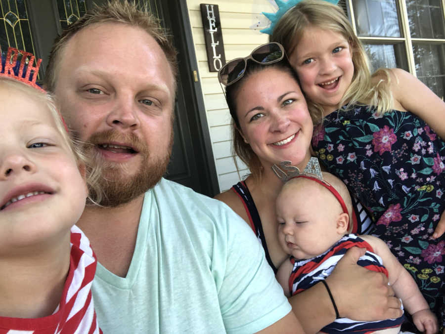 Mother sits smiling in front of front door of home with husband and three kids