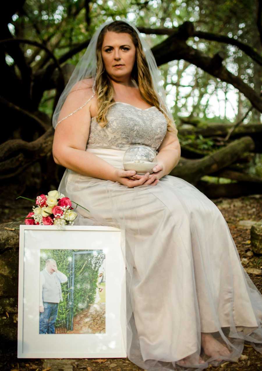 Woman in wedding dress sits in woods holding husbands ashes beside picture of him